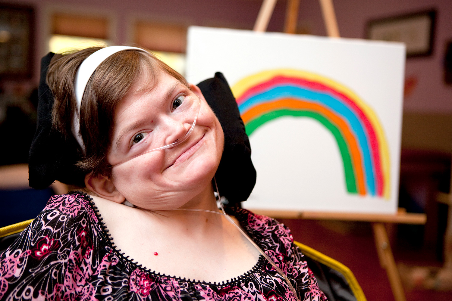 Woman wearing nasal canula smiles for the camera. Painting of rainbow is displayed on easel behind her.