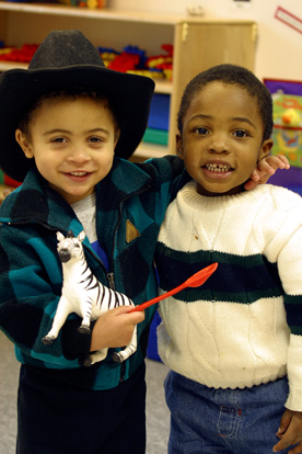 Young boy wears cowboy hat and holds toy zebra and spoon. Puts his arm around the shoulder of another young boy, as they both smile for the camera.
