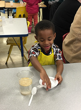 Child wearing yellow apron and holding a spoon works with ingredients in a small bowl