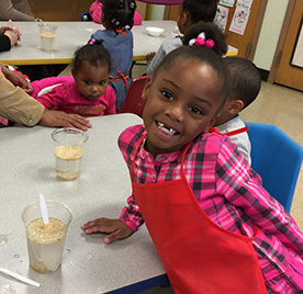 Young child wearing a red apron leans on a table and smiles for the camera