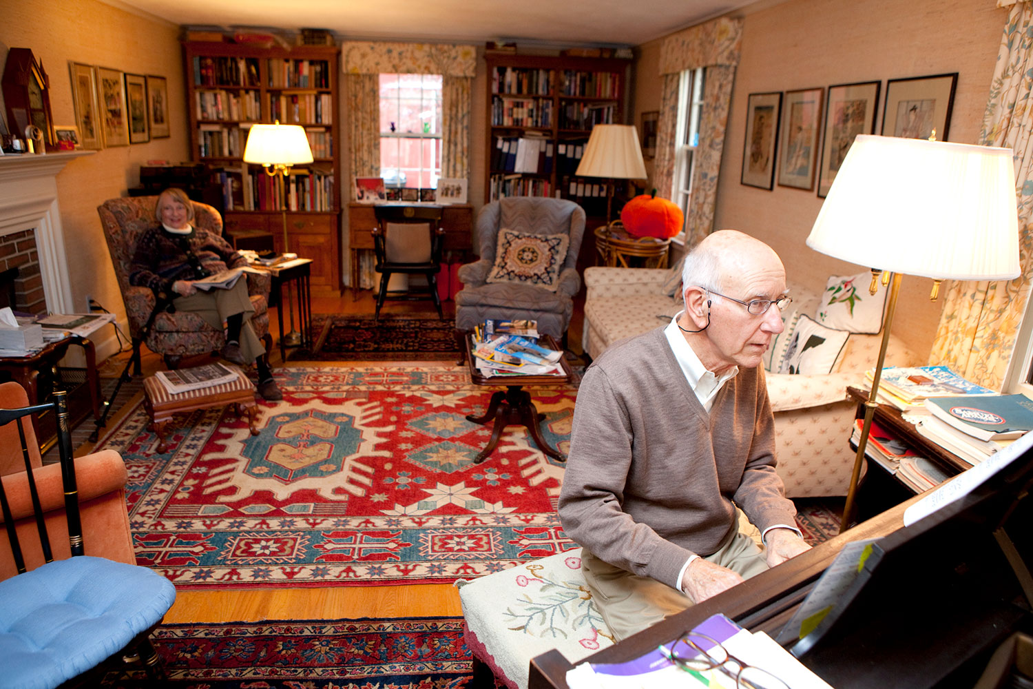 Older man plays piano in living room while seated woman looks on from across the room.