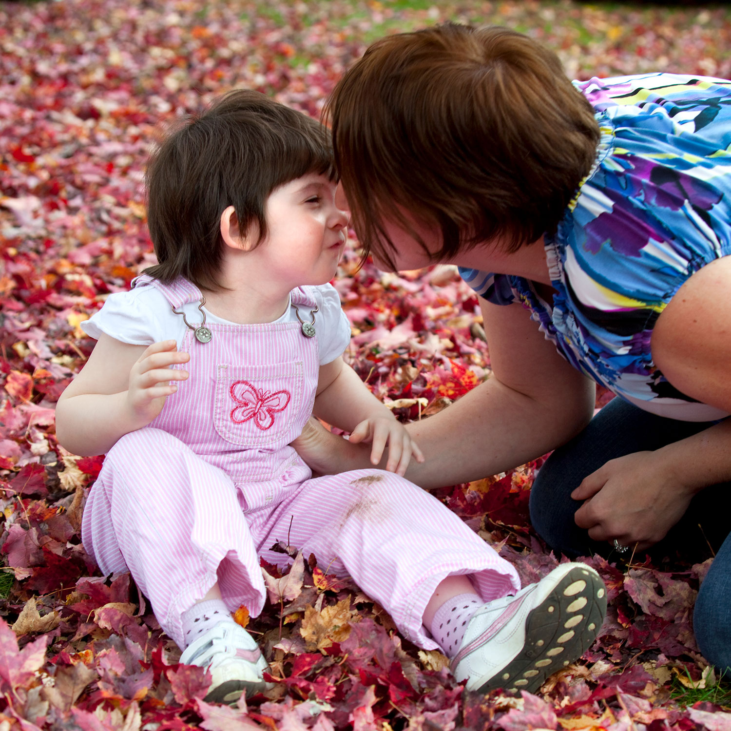 Toddler seated in autumn leaves outside reaches to kiss woman kneeling beside her