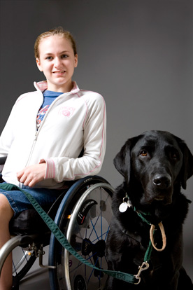 Young woman sits in wheel chair and smiles for the camera. Black Lab sits next to her and looks into the camera.